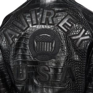 May include: Black leather jacket with a crocodile texture. The back of the jacket features the words "AVIREX USA" in large, raised letters. A circular emblem with an American flag design is centered above the text.