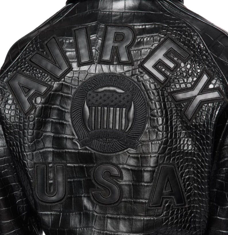 May include: Black leather jacket with a crocodile texture. The back of the jacket features the words "AVIREX USA" in large, raised letters. A circular emblem with an American flag design is centered above the text.