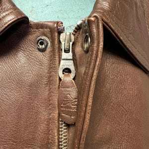 May include: Close-up of a brown leather jacket with a zipper. The zipper is silver and has the brand name "AVIREX" on it. The leather is a rich, dark brown color, and the jacket appears to be vintage.