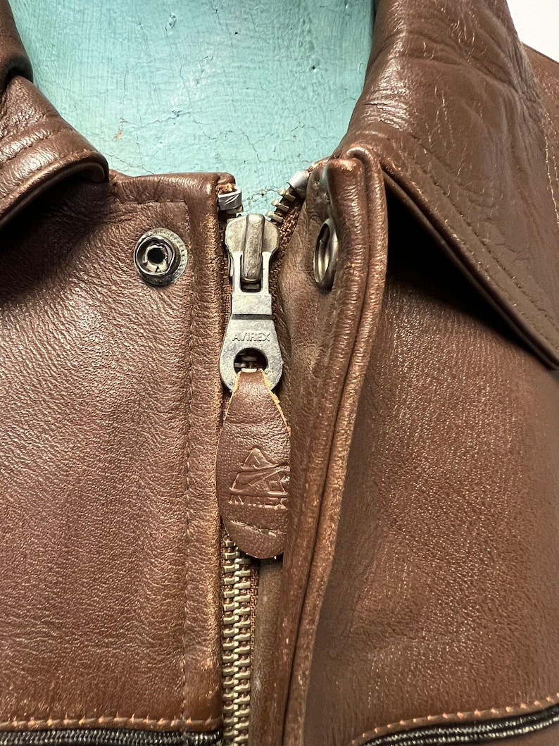 May include: Close-up of a brown leather jacket with a zipper. The zipper is silver and has the brand name "AVIREX" on it. The leather is a rich, dark brown color, and the jacket appears to be vintage.