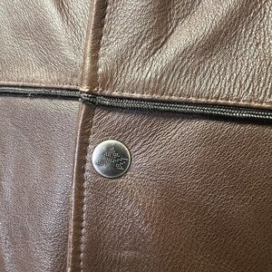 May include: Close-up of a brown leather jacket with a silver button. The jacket has a textured surface and a black zipper detail. The button features a geometric design.