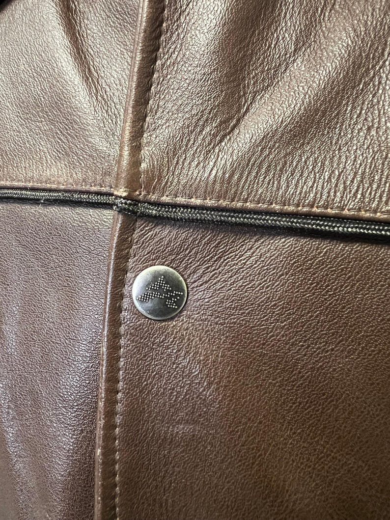 May include: Close-up of a brown leather jacket with a silver button. The jacket has a textured surface and a black zipper detail. The button features a geometric design.