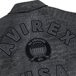 May include: Close-up of a gray jacket with a crocodile skin texture. The jacket features the word "AVIREX" in large, gray letters, along with a patch that reads "ALL AMERICANS ESTABLISHED 1975". A small logo is visible on the collar.