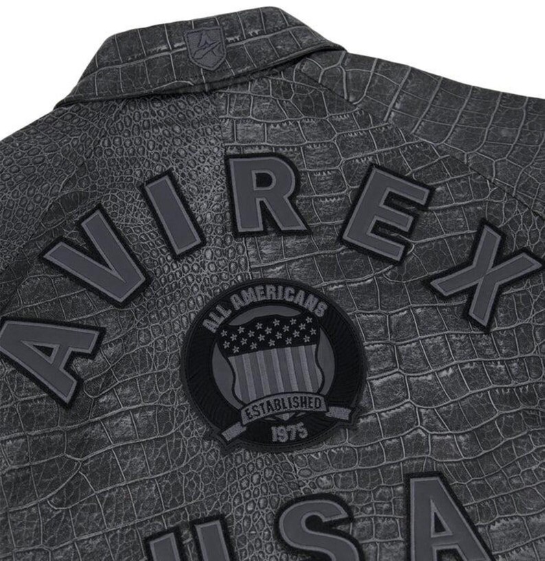 May include: Close-up of a gray jacket with a crocodile skin texture. The jacket features the word "AVIREX" in large, gray letters, along with a patch that reads "ALL AMERICANS ESTABLISHED 1975". A small logo is visible on the collar.