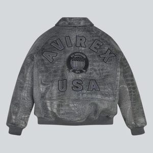 Puede incluir: Chaqueta de cuero gris con textura de cocodrilo. La parte trasera de la chaqueta presenta la palabra "AVIREX" arqueada en la parte superior, con "USA" debajo de un emblema circular. Cuello, pu&ntilde;os y cintura de canal&eacute;.