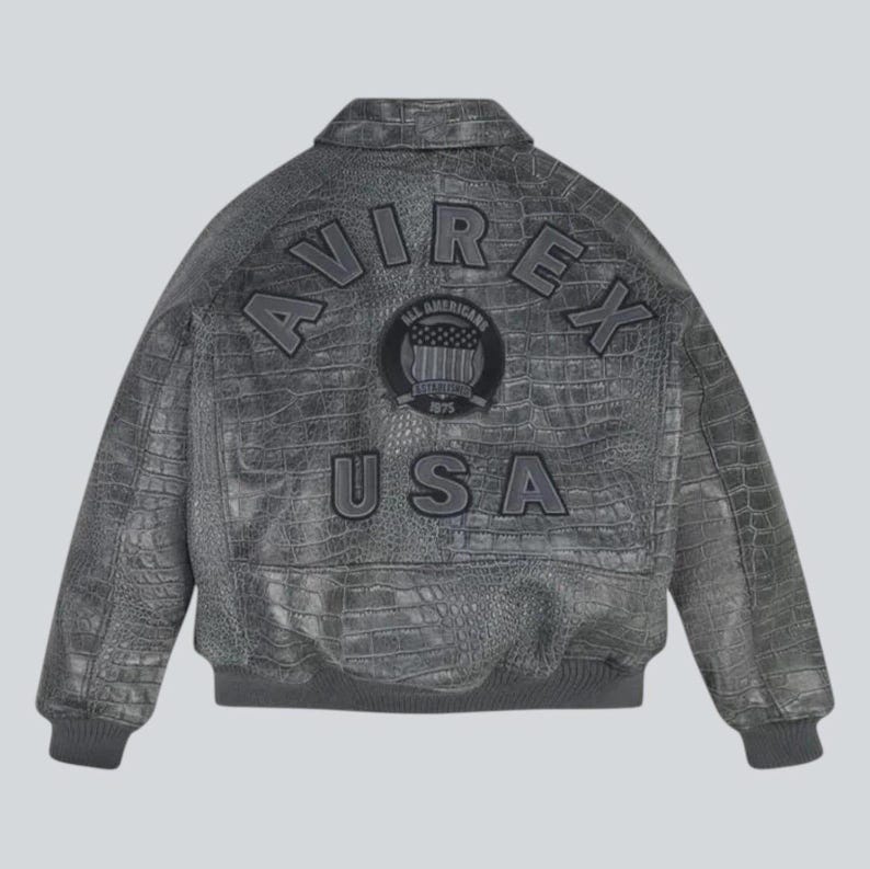 Puede incluir: Chaqueta de cuero gris con textura de cocodrilo. La parte trasera de la chaqueta presenta la palabra "AVIREX" arqueada en la parte superior, con "USA" debajo de un emblema circular. Cuello, pu&ntilde;os y cintura de canal&eacute;.