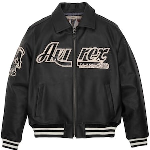 May include: Black leather jacket with a zip-up front, collar, and white and black striped cuffs and hem. The jacket features the text "Avi Rex BLACKACES" in white and a patch with a graphic and the number "52".