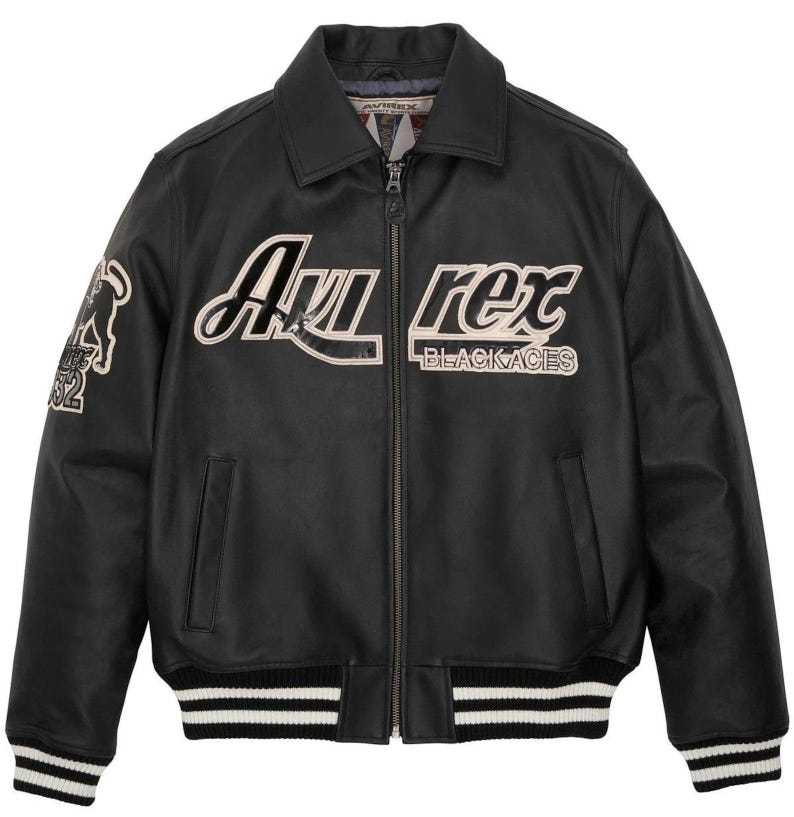 May include: Black leather jacket with a zip-up front, collar, and white and black striped cuffs and hem. The jacket features the text "Avi Rex BLACKACES" in white and a patch with a graphic and the number "52".