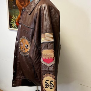 May include: A brown leather jacket with multiple patches, including an American flag and a patch with the number 55. The jacket has a button-down front and a collar. The patches are in shades of gold, red, and black.