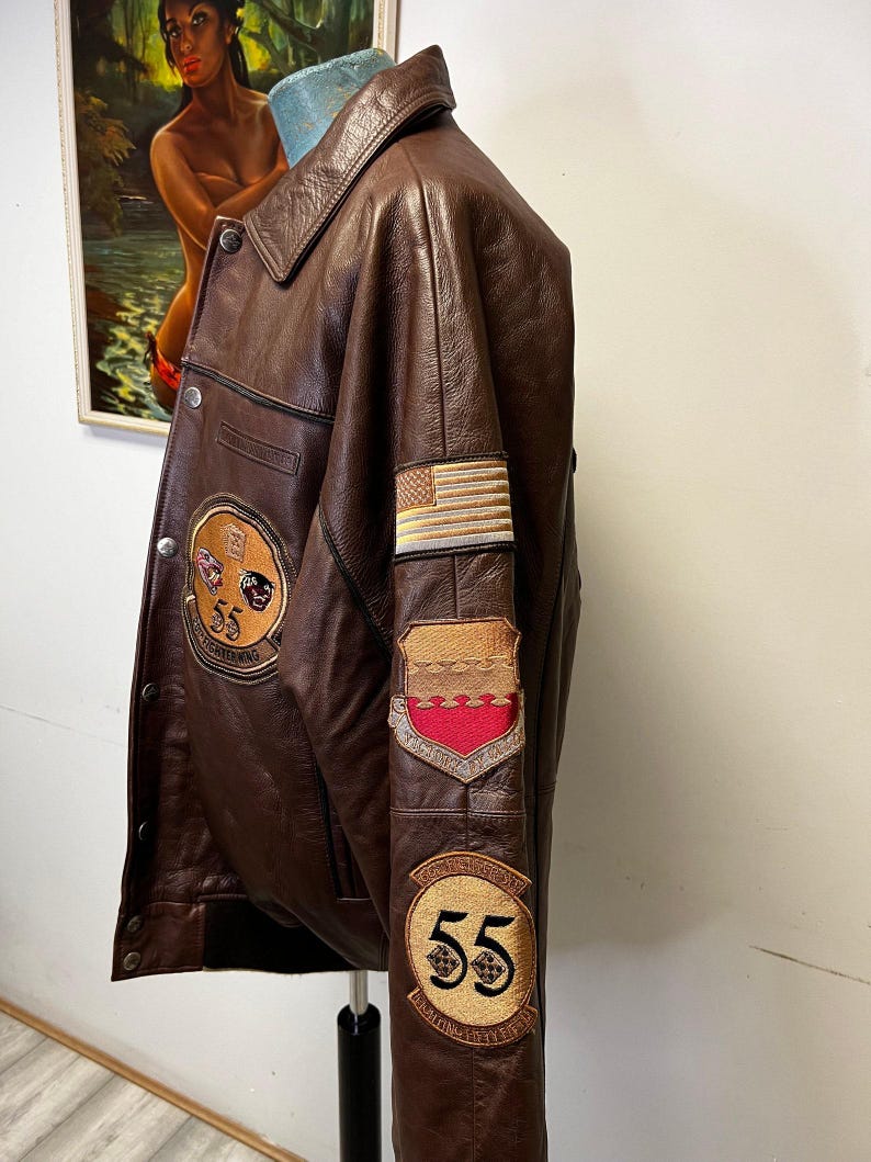 May include: A brown leather jacket with multiple patches, including an American flag and a patch with the number 55. The jacket has a button-down front and a collar. The patches are in shades of gold, red, and black.