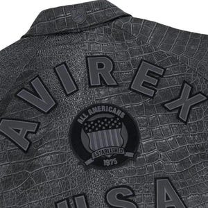 Puede incluir: Primer plano de una chaqueta gris con textura de piel de cocodrilo. La chaqueta presenta la palabra "AVIREX" en letras grandes y grises en relieve. Un parche circular dice "ALL AMERICANS ESTABLISHED 1975" con un dise&ntilde;o de bandera estadounidense.