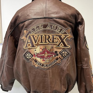 May include: Brown leather bomber jacket with a large embroidered patch on the back. The patch features the text "ALL ACES AVIREX 55th NO JOKERS" with playing card and airplane imagery. The jacket has a collared neckline and a ribbed hem.