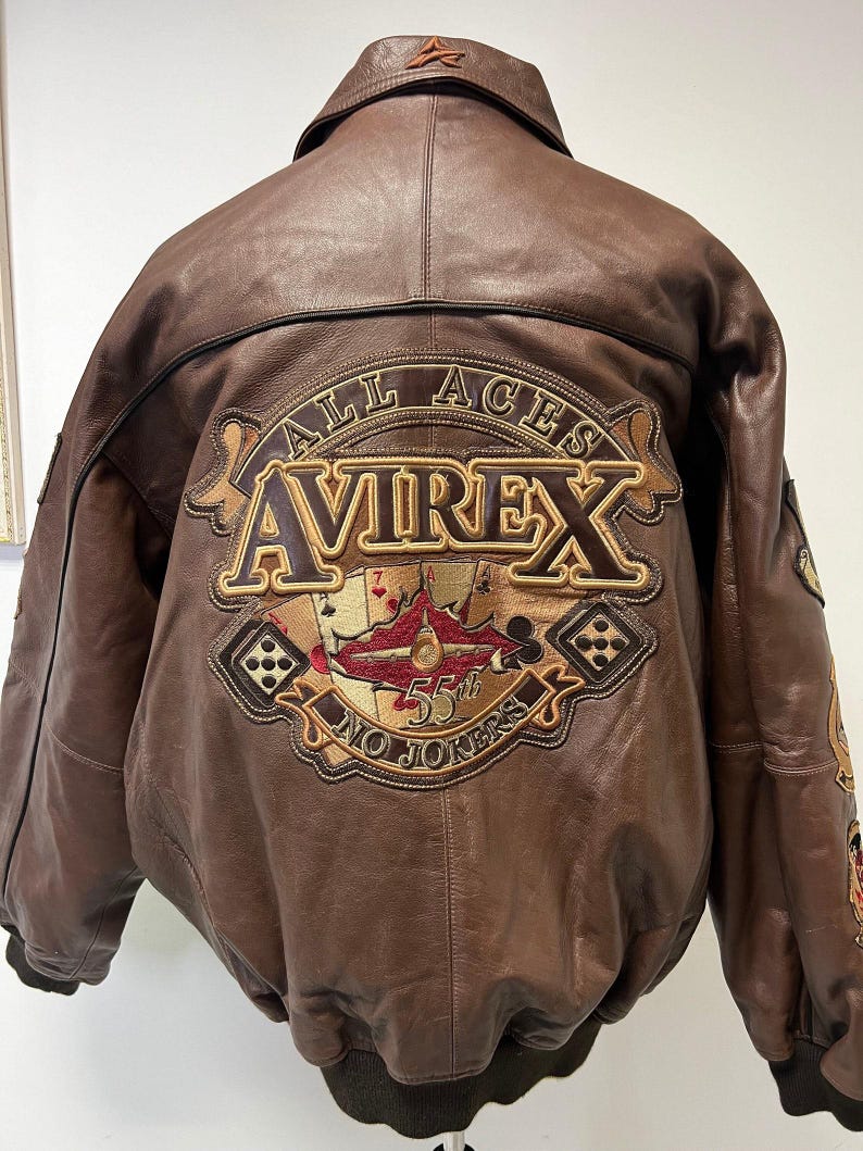 May include: Brown leather bomber jacket with a large embroidered patch on the back. The patch features the text "ALL ACES AVIREX 55th NO JOKERS" with playing card and airplane imagery. The jacket has a collared neckline and a ribbed hem.
