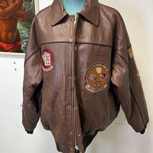 May include: Brown leather bomber jacket with a button-up front, collar, and multiple patches. Patches include playing card suits and text that reads "20th Fighter Wing" and "Fighter Sq". The jacket has a zipper closure and a ribbed hem.