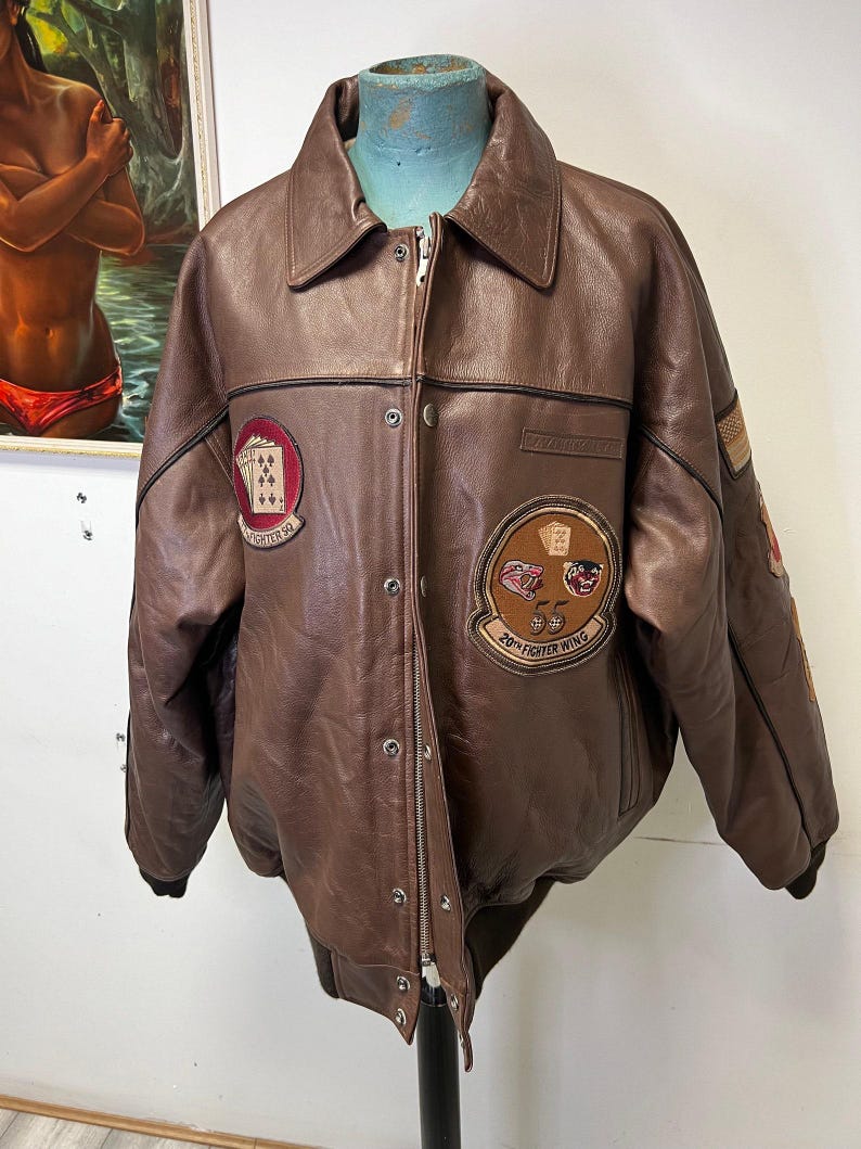 May include: Brown leather bomber jacket with a button-up front, collar, and multiple patches. Patches include playing card suits and text that reads "20th Fighter Wing" and "Fighter Sq". The jacket has a zipper closure and a ribbed hem.