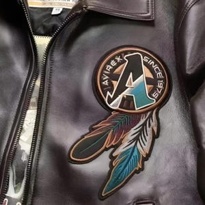 Op de afbeelding: Close-up van een zwarte leren jas met ritssluiting. De jas heeft een cirkelvormige patch met de tekst "AVIREX SINCE 1975" en een gestileerde "A", plus twee geborduurde veren in bruin, blauwgroen en blauw.