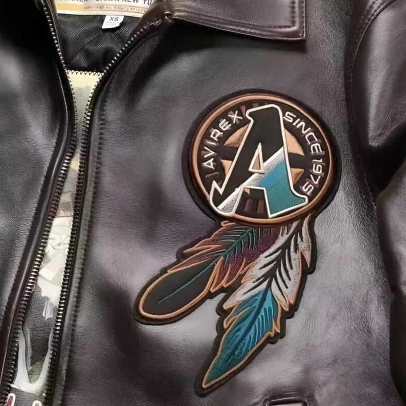 Op de afbeelding: Close-up van een zwarte leren jas met ritssluiting. De jas heeft een cirkelvormige patch met de tekst "AVIREX SINCE 1975" en een gestileerde "A", plus twee geborduurde veren in bruin, blauwgroen en blauw.