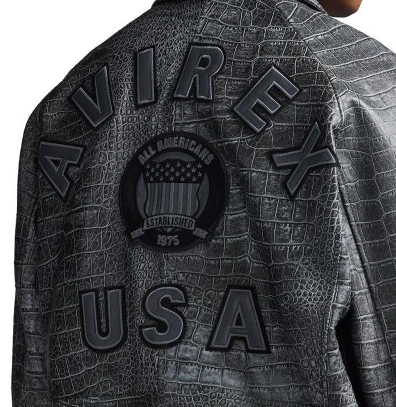 May include: A dark gray jacket with a crocodile skin texture. The back of the jacket features the words "AIR," "REX," and "USA" in raised, dark gray lettering. A circular patch with "ALL AMERICANS" and "ESTABLISHED 1975" is also visible.