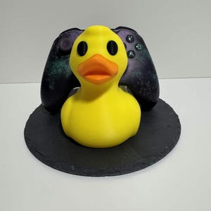 Könnte beinhalten: Eine quietschgelbe Gummiente mit schwarzen Augen und einem orangefarbenen Schnabel sitzt auf einer schwarzen, runden Schieferplatte. Hinter der Ente befindet sich ein schwarzer und violetter Videospiel-Controller. Die Ente ist ein lustiger, neuartiger Artikel.
