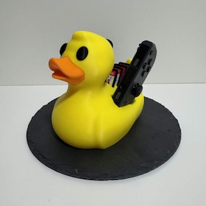 Könnte beinhalten: Eine gelbe Gummiente mit schwarzen Augen und einem orangefarbenen Schnabel, die einen schwarzen Gamecontroller und Spielkassetten hält. Die Ente steht auf einer schwarzen, runden, strukturierten Oberfläche. Ein lustiges und einzigartiges Gaming-Zubehör.