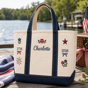 Op de afbeelding: Crèmekleurige draagtas met marineblauwe accenten en handvatten. De tas heeft geborduurde bloemen- en sterontwerpen, de naam "Charlotte" en de cijfers "1776" en "2026". De tas staat op een houten steiger bij een meer.