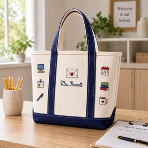 Op de afbeelding: Een crèmekleurige canvas tas met marineblauwe randen en hengsels. De voorzijde is voorzien van geborduurde iconen waaronder een computer, ID-badge, pen, envelop met appel, kalender, boeken en een mok, met de tekst Mrs. Bennett in blauw schrift.
