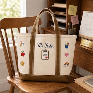 May include: A cream-colored canvas tote bag with brown trim and handles. The bag features embroidered icons including paper clips, an apple, a bell, a pencil cup, a heart, and a rainbow. The text Ms. Parker is embroidered above a clipboard icon.