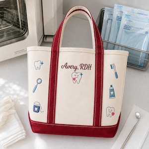 May include: A cream-colored canvas tote bag with dark red trim and handles. The bag features embroidered dental-themed icons including teeth, a toothbrush, toothpaste, and a mirror. The text Avery, RDH is embroidered in dark red script.