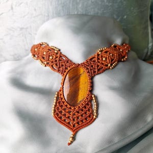Könnte beinhalten: Handgefertigte Makramee-Halskette mit einem großen ovalen Tigerauge-Stein-Anhänger. Die Halskette ist aus orange-braunem Kordel gefertigt und mit kleinen goldfarbenen Perlen verziert. Das aufwendige Makramee-Design verleiht einen Boho-Touch.
