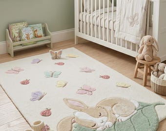 May include: A rectangular area rug in cream featuring a large, tufted, sleeping tan rabbit resting on a sage green leaf. The rug is decorated with small, colorful, tufted butterflies and ladybugs in shades of pink, yellow, and purple.
