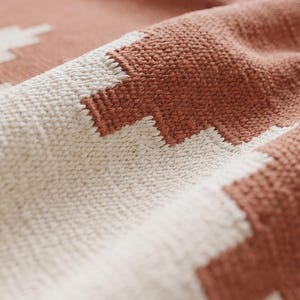 May include: A woven textile featuring a repeating geometric pattern in terracotta orange and off-white. The fabric displays a distinct textured weave with visible interlaced threads, creating a soft, folded appearance across the surface.