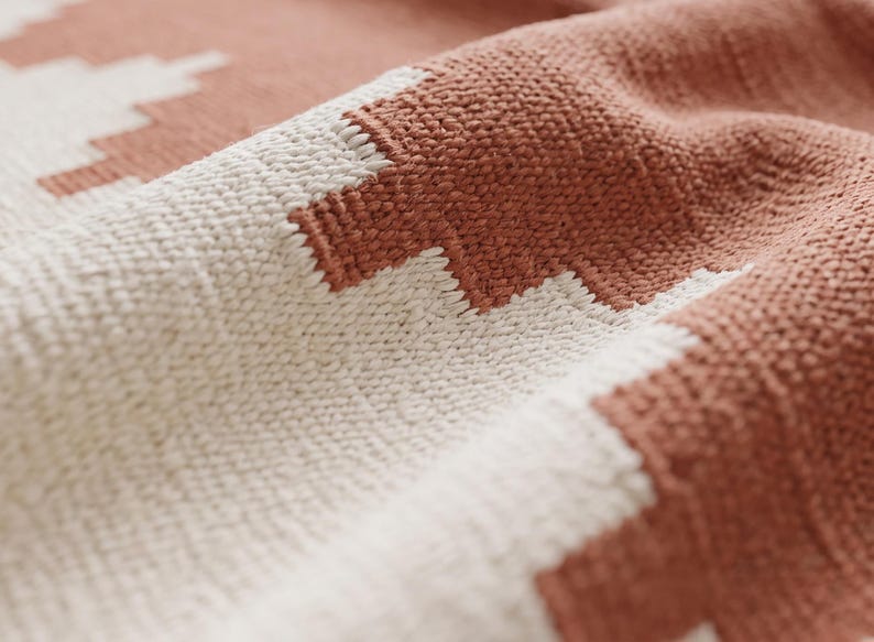 May include: A woven textile featuring a repeating geometric pattern in terracotta orange and off-white. The fabric displays a distinct textured weave with visible interlaced threads, creating a soft, folded appearance across the surface.