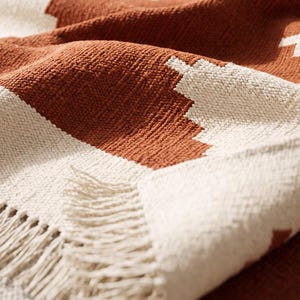 May include: A woven throw blanket featuring a geometric pattern in burnt orange and off-white. The fabric has a textured, nubby surface and is finished with a fringe edge consisting of loose, hanging threads along the bottom border.