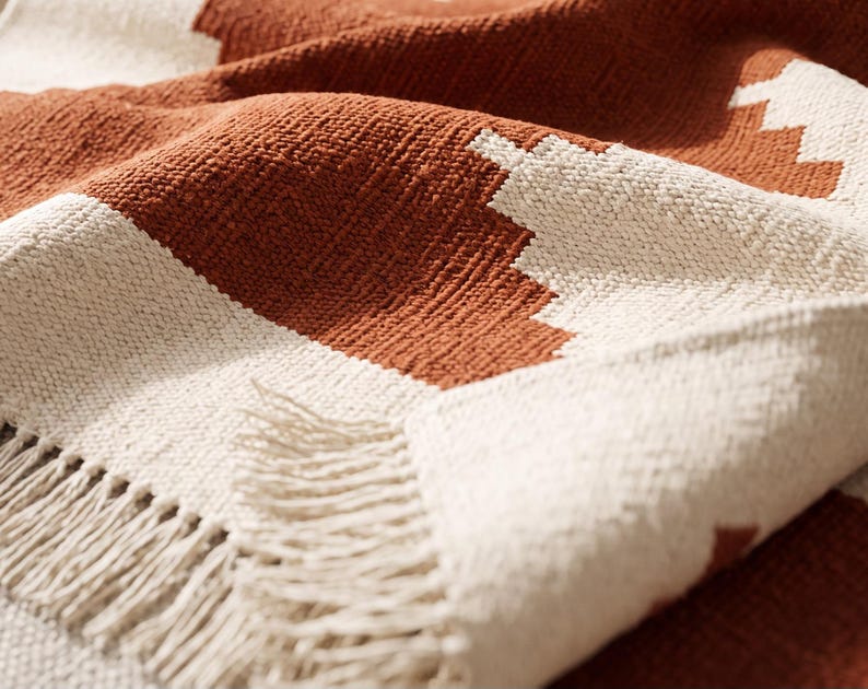 May include: A woven throw blanket featuring a geometric pattern in burnt orange and off-white. The fabric has a textured, nubby surface and is finished with a fringe edge consisting of loose, hanging threads along the bottom border.