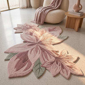 May include: A large, floral-shaped rug in shades of pink and green. The rug features a detailed design of overlapping petals and leaves, creating a three-dimensional effect. The rug is placed on a light-coloured floor, with a chair and side table in the background.