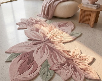May include: A large, floral-shaped rug in shades of pink and green. The rug features a detailed design of overlapping petals and leaves, creating a three-dimensional effect. The rug is placed on a light-colored floor, with a chair and side table in the background.