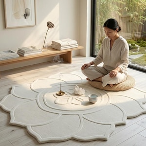 May include: A cream-colored, lotus flower-shaped rug with a raised design. A woman sits on a round, woven cushion on the rug. A small bowl, a crystal, and a brass incense holder are on the rug.