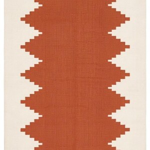 May include: A rectangular area rug featuring a central terracotta-colored geometric pattern with jagged, stepped edges against a solid off-white background. The rug has a flat-woven texture and short, straight fringe along the top and bottom edges.