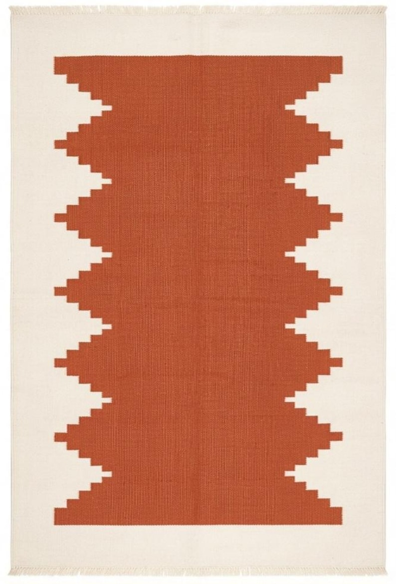 May include: A rectangular area rug featuring a central terracotta-colored geometric pattern with jagged, stepped edges against a solid off-white background. The rug has a flat-woven texture and short, straight fringe along the top and bottom edges.
