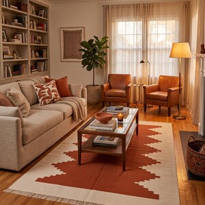 May include: A living room featuring a beige sofa with patterned pillows, two brown leather armchairs, and a wooden coffee table with a glass top. A large area rug with a geometric rust-colored pattern and fringe sits on the hardwood floor.