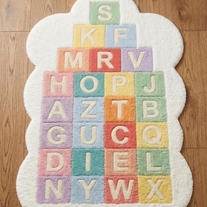 May include: A white, cloud-shaped rug with a colorful alphabet block design. Each block features a letter in a pastel color, creating a playful and educational accent for a child's room. The rug is made of soft, plush material.