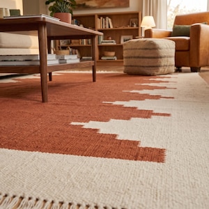 May include: A rectangular area rug featuring a two-tone geometric pattern with a large terracotta-colored center and cream-colored borders. The rug has a woven texture and is finished with short, cream-colored fringe along the short edge.