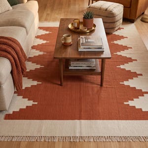 May include: A rectangular area rug featuring a geometric stepped pattern in burnt orange and cream. The rug has a flat-woven texture and is finished with short, cream-colored fringe along the shorter edges, covering the floor space.