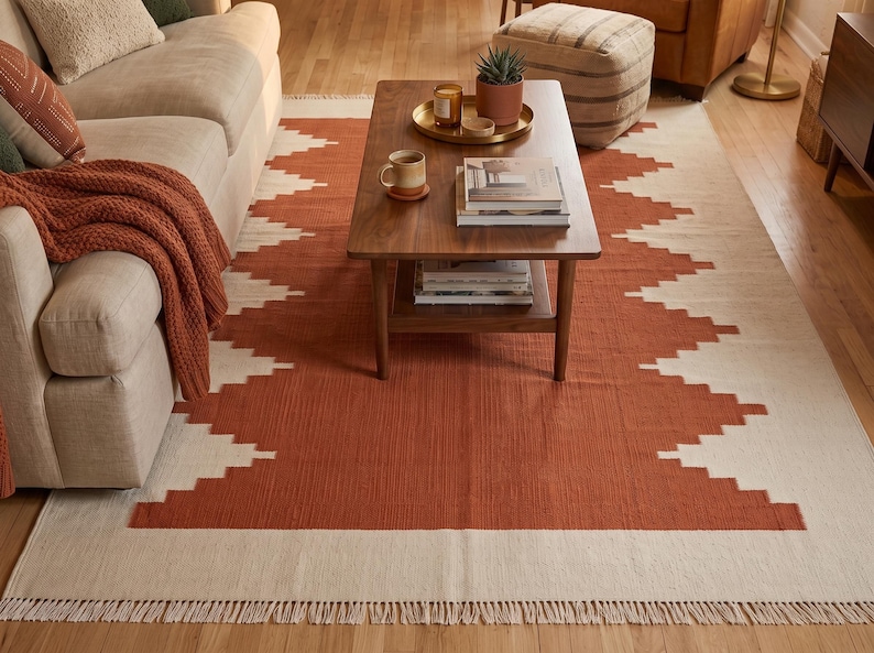 May include: A rectangular area rug featuring a geometric stepped pattern in burnt orange and cream. The rug has a flat-woven texture and is finished with short, cream-colored fringe along the shorter edges, covering the floor space.