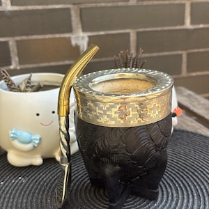 Può includere: Un contenitore per mate in zucca intagliata di colore marrone scuro, caratterizzato da un ampio bordo in metallo dorato con un complesso motivo geometrico. Una bombilla dorata abbinata, con stelo intrecciato e base filtrante piatta, poggia contro.