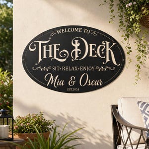 May include: An oval black metal sign with white lettering that reads "WELCOME TO THE DECK", "SIT RELAX ENJOY", "Mia & Oscar", and "EST.2024". The sign features decorative flourishes and is mounted on a wall, displaying a cut-out style design.