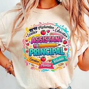 May include: Cream-colored t-shirt with a colorful graphic design. The design features the words "Assistant Principal" in large letters, surrounded by other words like "Leader" and "Educator", along with hearts, apples, and a pencil.