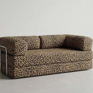 Könnte beinhalten: Zweisitzer-Sofa mit Leopardenmuster. Das Sofa hat ein niedriges Profil mit abgerundeten Armlehnen und einer rechteckigen Sitzfläche. Der Rahmen ist mit silberfarbenen Metallstützen versehen. Die Polsterung weist ein braunes und schwarzes Fleckenmuster auf.