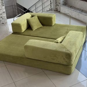 Op de afbeelding: Een modulaire, olijfgroene corduroy bank met twee secties en twee bijpassende vierkante sierkussens. De bank heeft een laag profiel en een modern design, geschikt voor een woonkamer of loungeruimte. De corduroy stof voegt textuur toe.