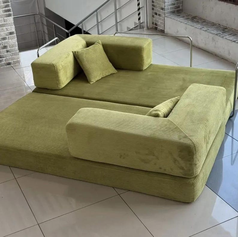 Op de afbeelding: Een modulaire, olijfgroene corduroy bank met twee secties en twee bijpassende vierkante sierkussens. De bank heeft een laag profiel en een modern design, geschikt voor een woonkamer of loungeruimte. De corduroy stof voegt textuur toe.